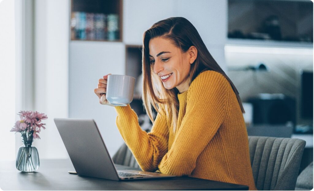 Junge Frau mit gelbem Pullover sitzt mit einer Tasse am Laptop und lächelt. Sie ist Sprachpartnerin bei Little World und nimmt gerade an einem Gespräch teil.